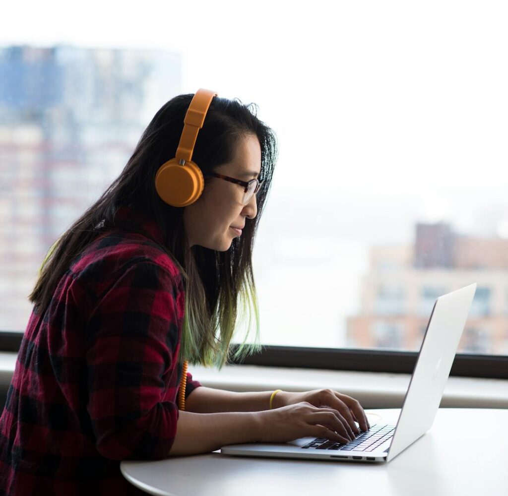 Asian woman with headphones using laptop for remote work in a city setting.